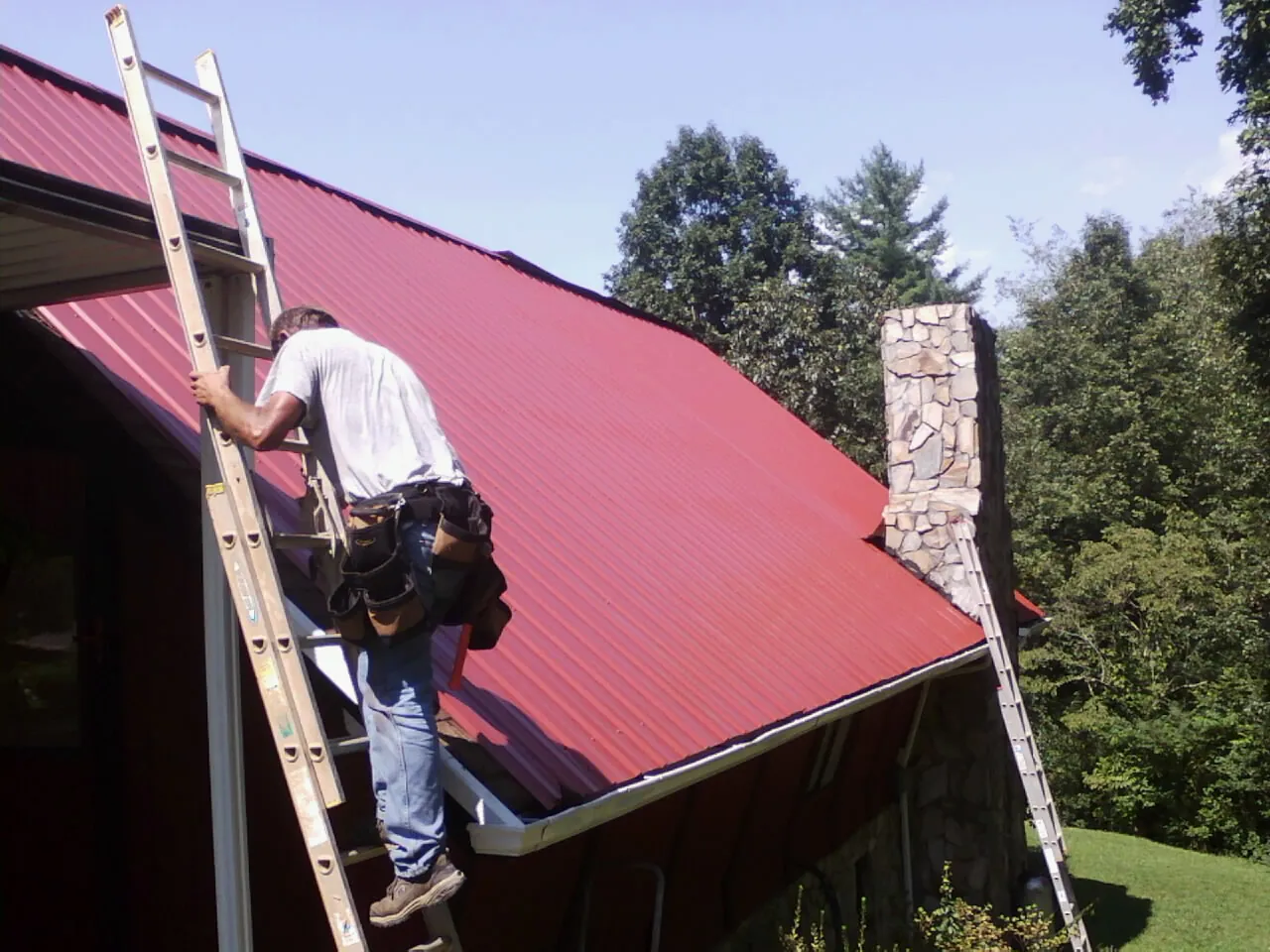 Roofer on a ladder beside a red metal roof and stone chimney — Soffit & Fascia Repair by Roofing Brothers in Koppel